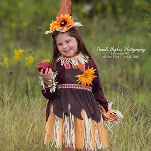 May include: A child in a handmade scarecrow costume, holding a red apple. The costume includes a brown hat with a sunflower, a burgundy top, and a brown and orange skirt with fringe. The outfit is set in a grassy field.