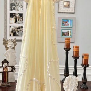 May include: A vintage-style, pale yellow gown with intricate lace detailing. The gown features short sleeves, a high neckline, and delicate embellishments. A matching bonnet with lace trim is also present. The words "Royal Gown Back" are written in script.