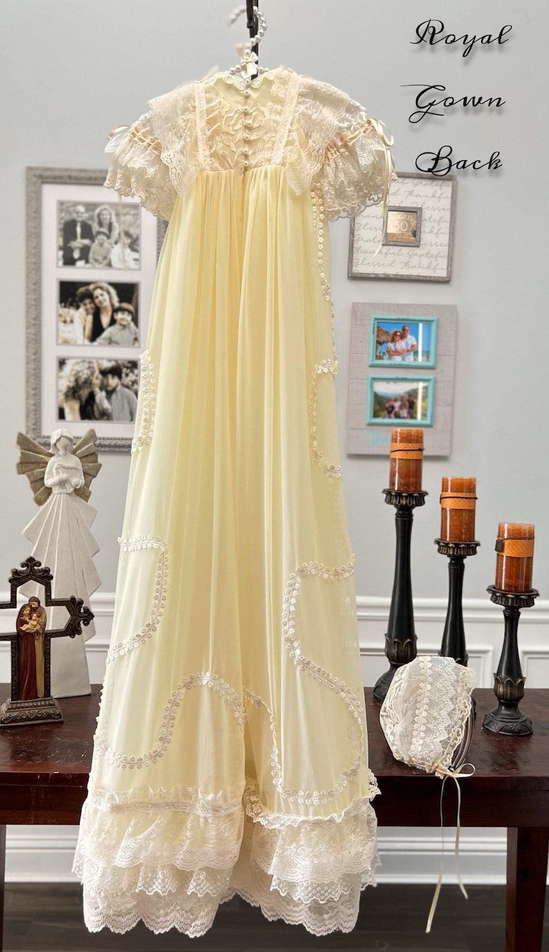 May include: A vintage-style, pale yellow gown with intricate lace detailing. The gown features short sleeves, a high neckline, and delicate embellishments. A matching bonnet with lace trim is also present. The words "Royal Gown Back" are written in script.