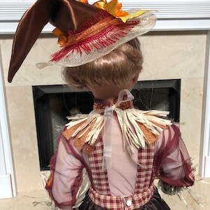 May include: A decorative scarecrow doll wearing a brown satin hat with orange and yellow fall leaves. The doll has a plaid bodice, sheer sleeves, and a brown skirt. The doll is decorated with straw and glitter accents.