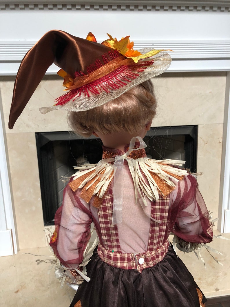 May include: A decorative scarecrow doll wearing a brown satin hat with orange and yellow fall leaves. The doll has a plaid bodice, sheer sleeves, and a brown skirt. The doll is decorated with straw and glitter accents.