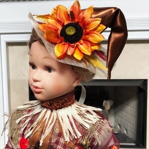 May include: A doll dressed as a scarecrow, wearing a brown hat with a large orange sunflower and a decorative collar. The scarecrow outfit includes a plaid shirt with red buttons and a straw-like collar, perfect for fall decor.