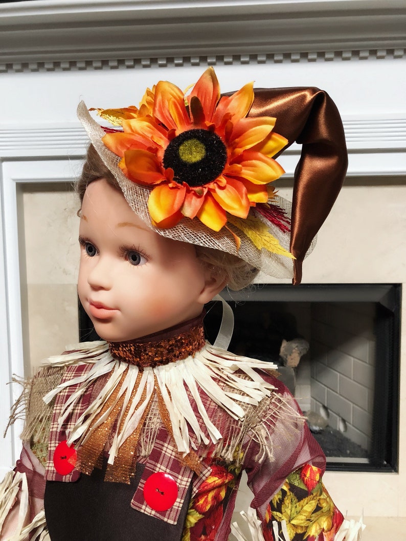 May include: A doll dressed as a scarecrow, wearing a brown hat with a large orange sunflower and a decorative collar. The scarecrow outfit includes a plaid shirt with red buttons and a straw-like collar, perfect for fall decor.