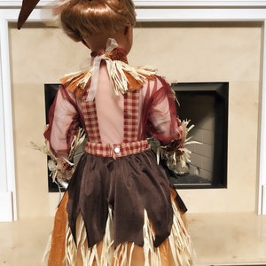 May include: A decorative scarecrow doll, viewed from the back, with a brown hat adorned with leaves. The doll wears a plaid shirt, a brown skirt with straw accents, and white boots. The outfit is in autumn colors.