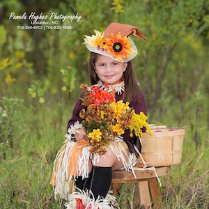 May include: A young girl dressed as a scarecrow, wearing a brown hat adorned with sunflowers and fall foliage. She holds a bouquet of autumn flowers and sits on a wooden stool. The outfit includes a fringed skirt and leg warmers.