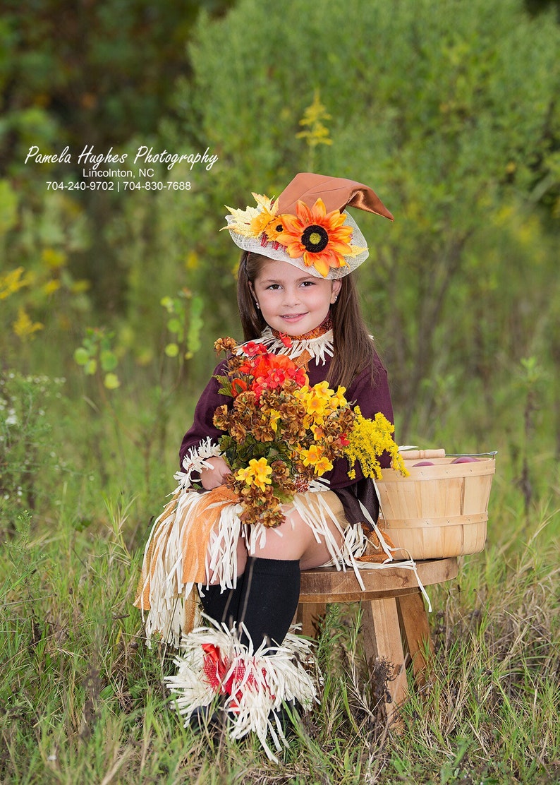 May include: A young girl dressed as a scarecrow, wearing a brown hat adorned with sunflowers and fall foliage. She holds a bouquet of autumn flowers and sits on a wooden stool. The outfit includes a fringed skirt and leg warmers.
