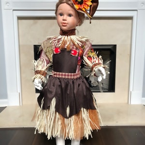 May include: A decorative doll dressed as a scarecrow, featuring a brown and orange color scheme. The doll wears a hat with a sunflower, a brown dress with straw accents, and white tights. The doll is standing in front of a fireplace.