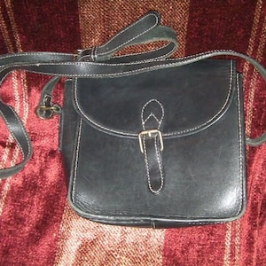 May include: A black leather crossbody bag with a flap closure and a buckle. The bag has a long adjustable strap and a small pocket on the front.