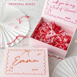 Könnte beinhalten: Rosa Brautjungfern-Angebotsboxen mit gewellten Kanten. Eine Box ist geöffnet und mit rotem und rosa Crinkle-Papier gefüllt. Der Deckel der offenen Box trägt den Text "Willst du meine... Brautjungfer sein?" Die andere Box trägt den Namen "Emma".