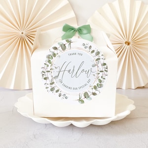 May include: White favour box with a green eucalyptus wreath and gold lettering that says "Thank You Harlow for Sharing Our Special Day".
