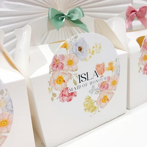 Puede incluir: Cajas de regalo blancas con pegatinas florales y lazos pastel. Las cajas tienen asas y están decoradas con una pegatina circular con flores de acuarela y el texto "ISLA MAID OF HONOUR". Un lazo verde menta adorna la parte superior de una caja.