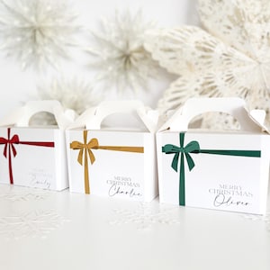 May include: Three white gift boxes with handles, each featuring a different coloured bow: red, gold, and green. The boxes are labelled "Merry Christmas" and a name in a script font. Decorative snowflakes and floral accents are in the background.