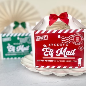 May include: Christmas-themed gift boxes, one red and one green, both with "Elf Mail" text. The red box has a red bow, the green box a green bow. The boxes are on a white scalloped plate, festive and ready for the holidays.