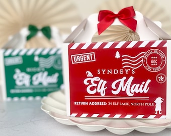 May include: White gift boxes with handles, decorated for Christmas. One box is red with "Elf Mail" text and a red bow. Another box is green with "Elf Mail" text and a green bow. The boxes are on a white scalloped plate.