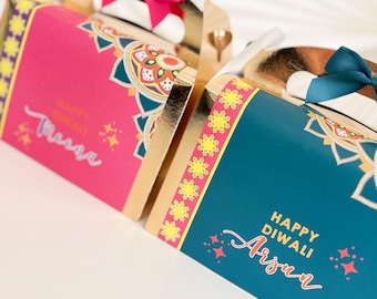 May include: Two decorative gift boxes, one pink and one teal, with gold accents and floral patterns. Each box has a bow and the words "Happy Diwali" and a name in script. The boxes are likely for a holiday.