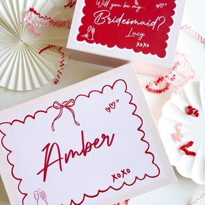 May include: Two pink gift boxes with red scalloped borders and red text. The top box says "Will you be my... Bridesmaid? Lucy xoxo" and the bottom box says "Amber xoxo" with a champagne glass illustration.