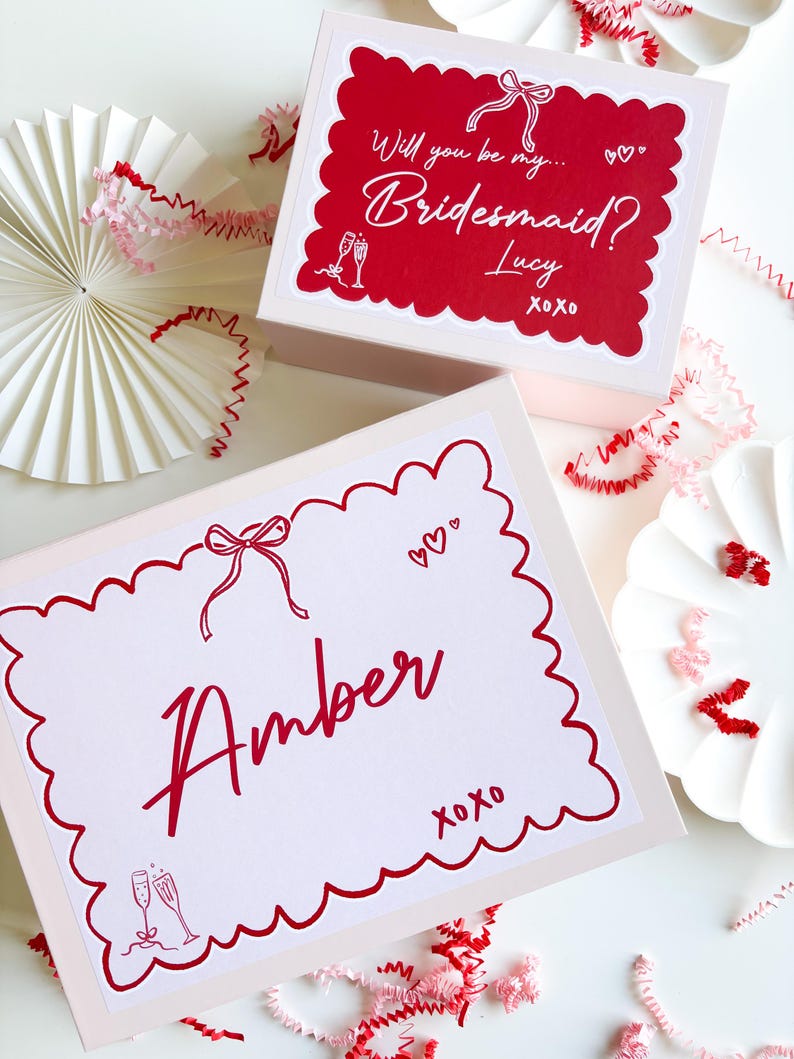 May include: Two pink gift boxes with red scalloped borders and red text. The top box says "Will you be my... Bridesmaid? Lucy xoxo" and the bottom box says "Amber xoxo" with a champagne glass illustration.