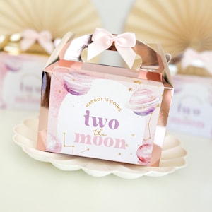 May include: A pink and gold favour box with a pink bow. The box has a watercolour design of planets and stars. The text on the box reads "Margot is going two the moon".