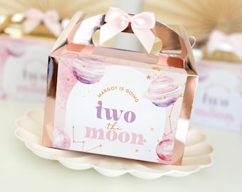 Puede incluir: Una caja de regalo rosa y dorada con un lazo rosa. La caja tiene un diseño de acuarela de planetas y estrellas. El texto en la caja dice "Margot is going two the moon".