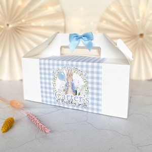 May include: White favor box with a blue gingham pattern and a blue bow. The box has a sticker with a cartoon bunny holding a blue balloon and the text "Thank you for coming to Carter's 1st Birthday Party."