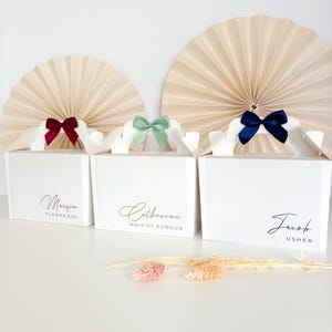 May include: Three white gift boxes with handles and bows. The boxes are labelled "Maggie Flower Girl", "Catherine Maid of Honour", and "Jacob Usher".