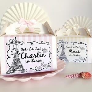 May include: Two pink and yellow striped favor boxes with a white Eiffel Tower design. The boxes have a pink and white bow on top and say "Ooh La La! Charlie in Paris" and "Ooh La La! Mari in Paris".
