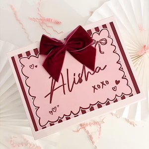 Könnte beinhalten: Eine rosa Geschenkbox mit einem gewellten Rand und dem Namen "Alisha" in burgunderfarbener Schrift. Eine burgunderfarbene Samtschleife sitzt auf der Box, mit kleinen Herzen und "xoxo" Akzenten. Die Box ist mit vertikalen Streifen verziert.