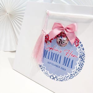 May include: White gift bag with a pink bow, tassel, and a decorative tag. The tag features the text "Sophia's Hen Mamma Mia September 2026" with a disco ball and floral accents. The bag has a white handle.