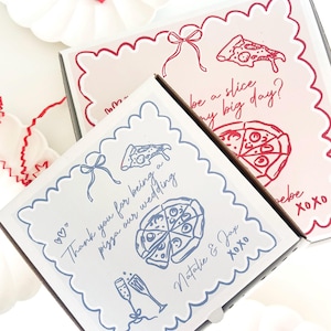 Può includere: Due scatole per pizza con bordi smerlati. Una scatola è bianca con illustrazioni blu e testo: "Thank you for being a pizza our wedding Natalie & Jax xoxo." L'altra scatola è rosa con illustrazioni rosse e testo: "be a slice my big day?"