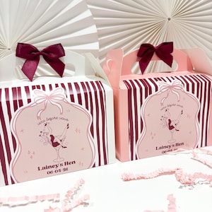 May include: Two gift boxes with burgundy and white stripes, each with a burgundy bow. The boxes have pink labels with a cartoon woman in a martini glass and the text "Lainey's Hen 06-04-26". A white fan-shaped design forms the background.
