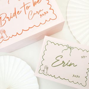 Könnte beinhalten: Zwei rosa Geschenkboxen mit gewellten Rändern und dekorativem Text. Auf einer Box steht "Bride to be Cara" in orangefarbener Schrift, auf der anderen "Erin" in grüner Schrift. Beide Boxen haben Schleifen- und Champagnerglas-Illustrationen.