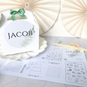 May include: A white favour box with a green bow and a sticker that says "JACOB" with a watercolour design of eucalyptus leaves. The box is sitting on a white plate next to a black and white printable activity sheet with games and puzzles for a wedding.