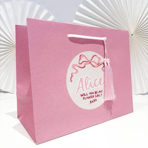May include: A pink gift bag with white rope handles and a pink tassel. The bag features a circular design with a bow and the name "Alice" and the text "Will you be my flower girl? xoxo".