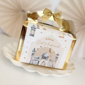 May include: A gold gift box with a white label featuring a mosque, stars, and a crescent moon. The label reads "Adam Eid Mubarak".