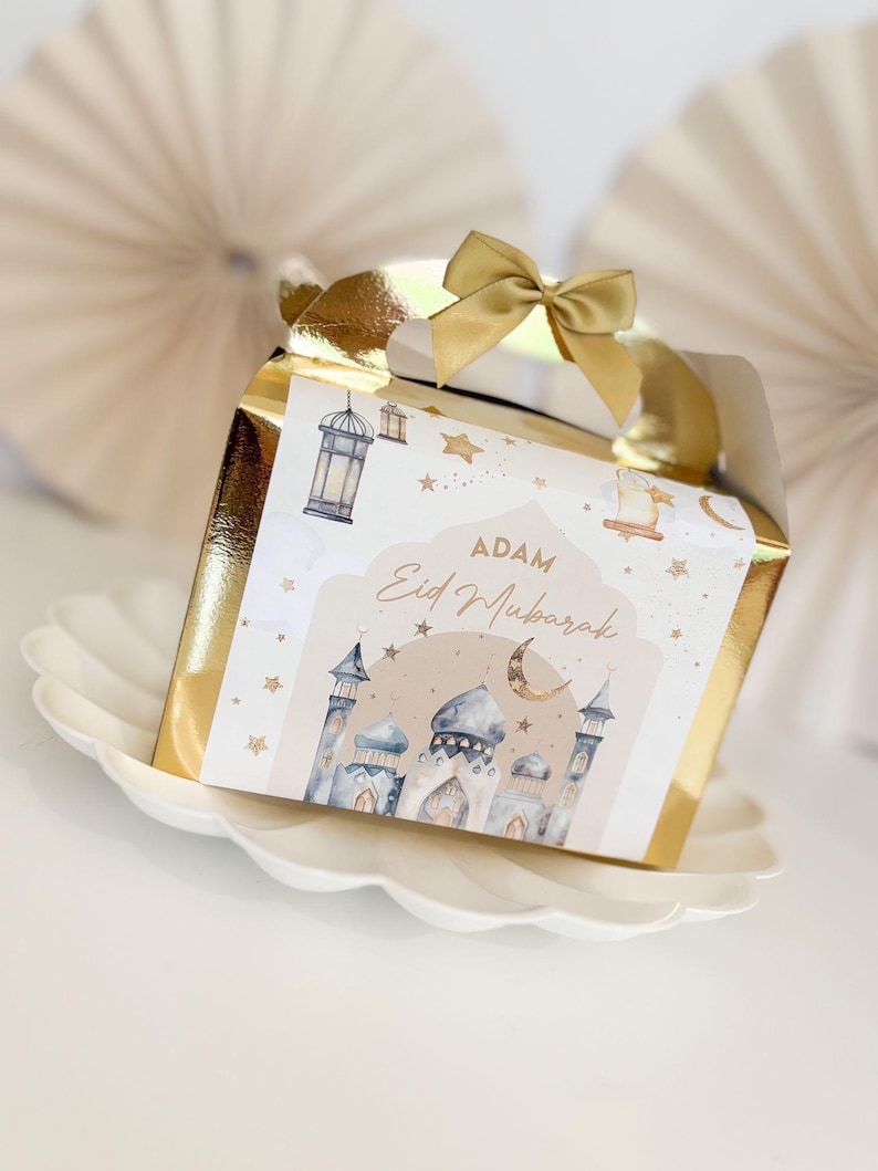 May include: A gold gift box with a white label featuring a mosque, stars, and a crescent moon. The label reads "Adam Eid Mubarak".
