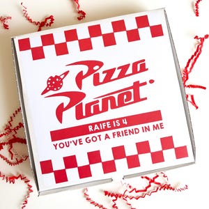 May include: A white pizza box with a red and white checkered border and the words "Pizza Planet" in red. The text "Raife is 4" and "You've got a friend in me" are also in red. Red paper shreds are scattered around the box.