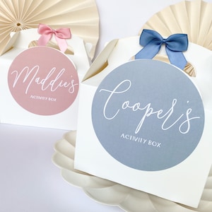 May include: Two white gift boxes with handles, one with a pink circle and the text "Maddie's Activity Box" and the other with a blue circle and the text "Cooper's Activity Box".