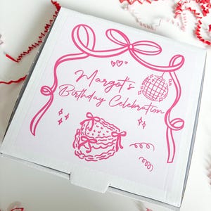 Può includere: Una scatola bianca con grafica rosa e testo che recita "Margot's Birthday Celebration". Il design include nastri, cuori, una palla da discoteca e un'illustrazione di torta. Coriandoli di carta rossi e rosa sono sparsi intorno alla scatola.