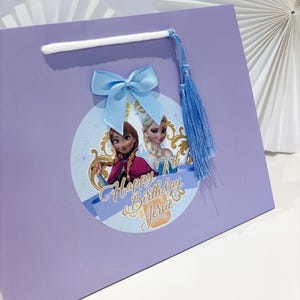 Puede incluir: Una bolsa de regalo lila con una imagen circular de dos princesas animadas y el texto "Happy Birthday Jerrie 7th". Tiene un lazo azul claro, una borla azul y un asa de cord&oacute;n blanco.