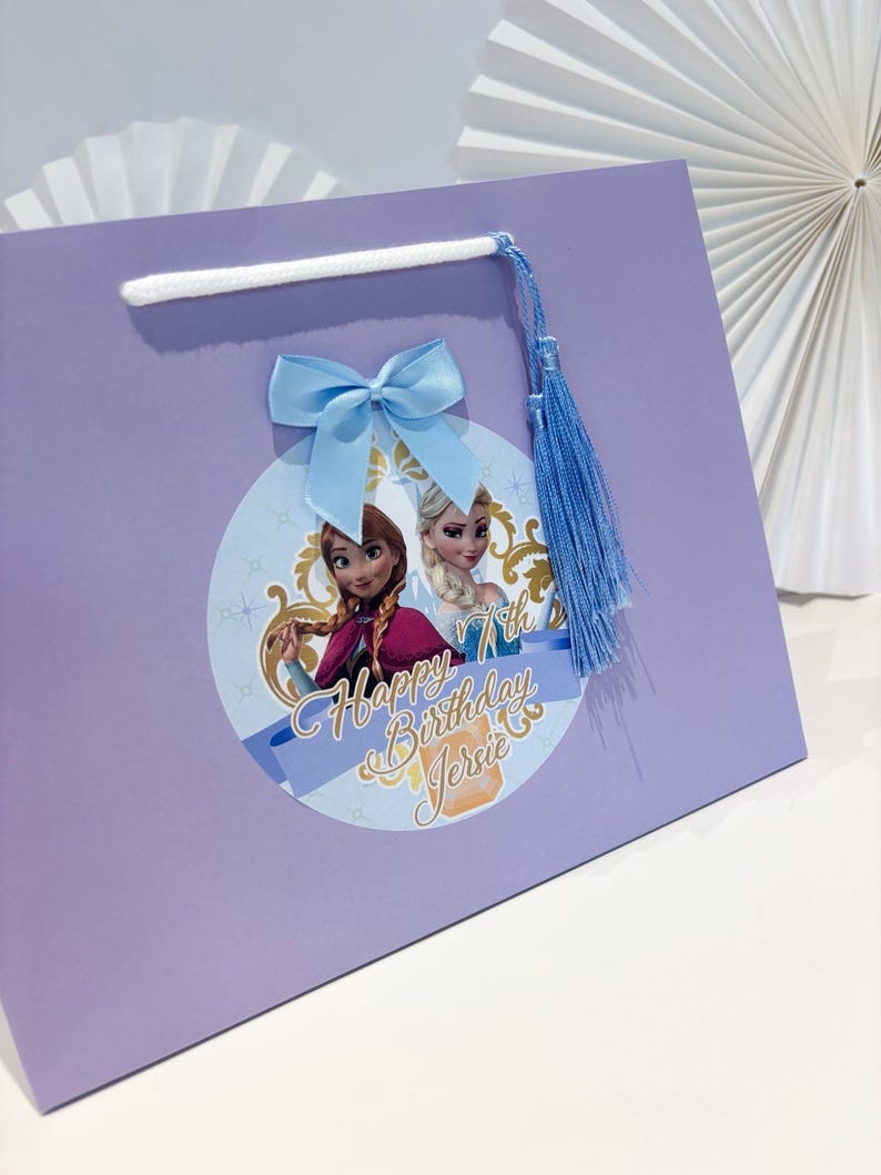 Puede incluir: Una bolsa de regalo lila con una imagen circular de dos princesas animadas y el texto "Happy Birthday Jerrie 7th". Tiene un lazo azul claro, una borla azul y un asa de cord&oacute;n blanco.