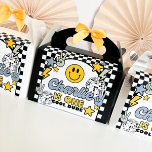 May include: Three black and white chequered party favour boxes with yellow ribbon handles. The boxes have a disco ball, peace sign, and smiley face design. The middle box has the text "Charlie is one cool dude".