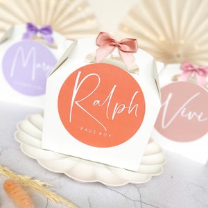 May include: Three white favour boxes with pink and purple ribbons. The boxes have round labels with names and titles. The labels read "Mary", "Ralph Page Boy", and "Vivi".