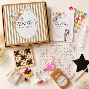 May include: A wedding activity box set featuring a striped cardboard box labelled Hallie's Activity Box, a wedding activity book, a snakes and ladders game board, a wooden tic-tac-toe set, a star-shaped chalkboard, crayons, a die, and stickers.