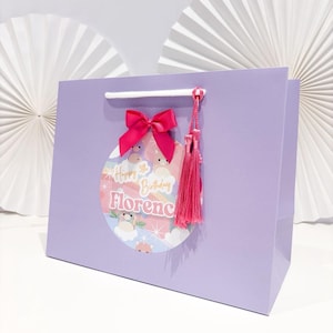May include: A lavender gift bag with a pink bow and tassel. The bag features a circular design with the text "Happy 5th Birthday Florence" and illustrations of clouds and bears. White paper fans are in the background.