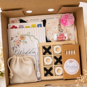 May include: A cardboard gift box containing a Snakes and Ladders game board, a wedding activity book, a wooden tic-tac-toe set with black and white pieces, a box of wedding crayons, a small beige drawstring bag, a bubble wand, and a Love Hearts sweet.
