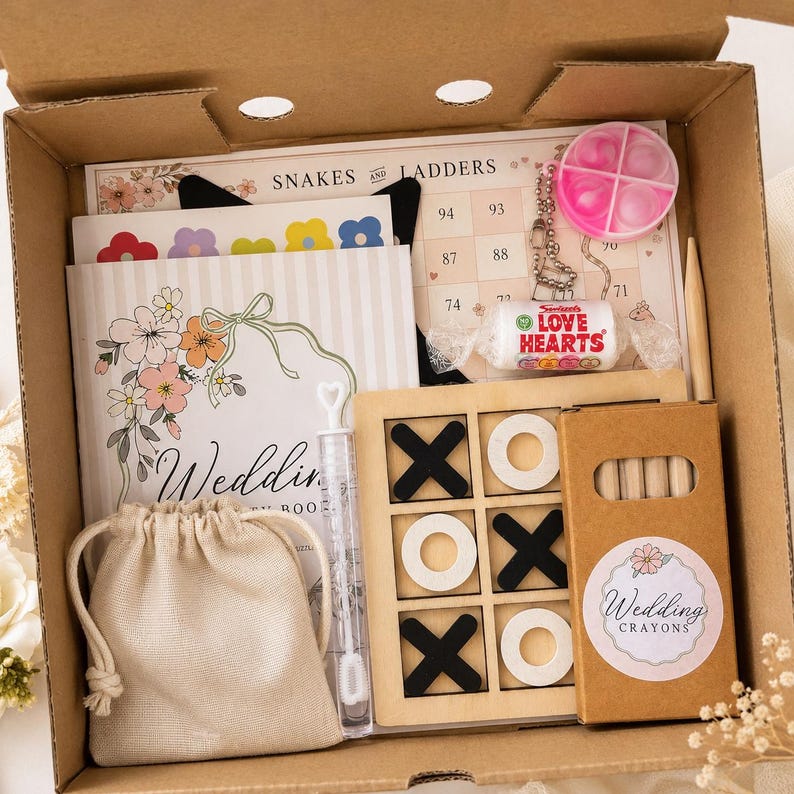 May include: A cardboard gift box containing a Snakes and Ladders game board, a wedding activity book, a wooden tic-tac-toe set with black and white pieces, a box of wedding crayons, a small beige drawstring bag, a bubble wand, and a Love Hearts sweet.