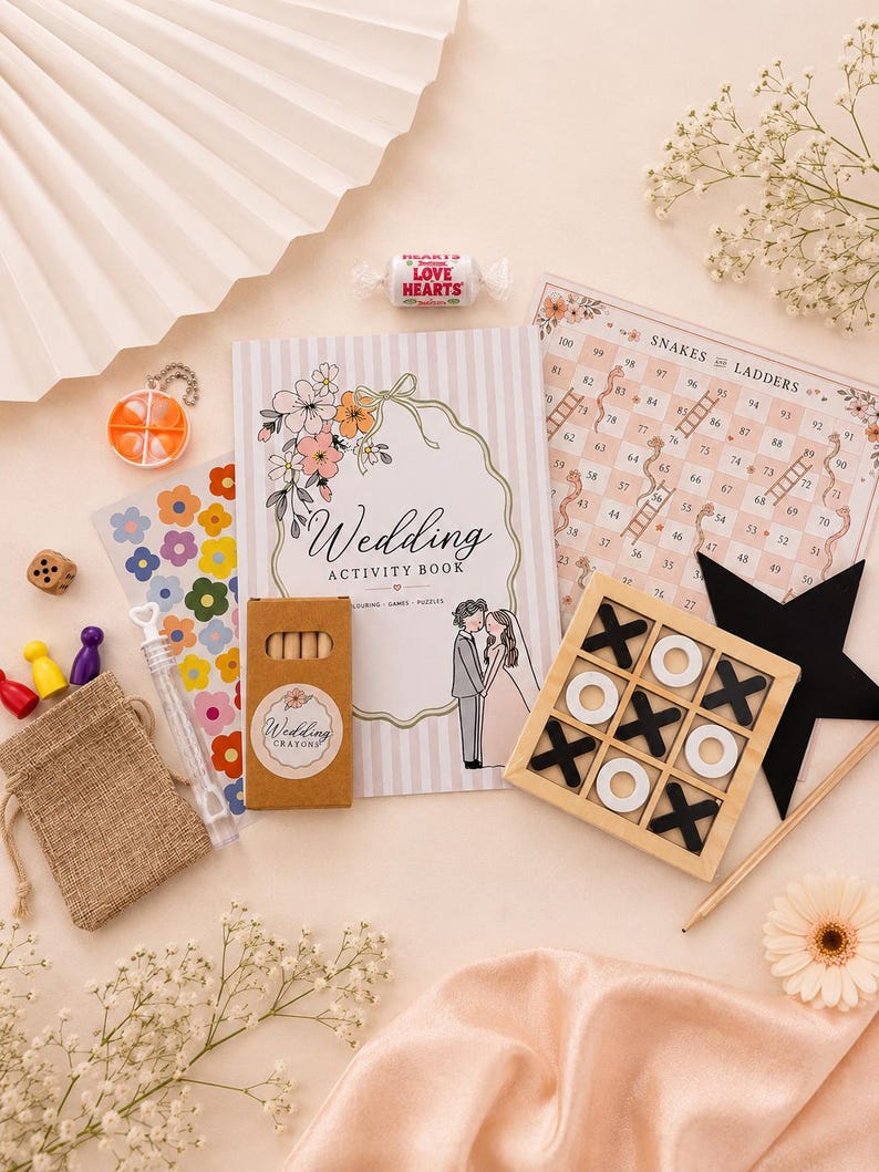 May include: A wedding activity book with a floral cover, a box of wedding crayons, a Snakes and Ladders game board, a wooden tic-tac-toe set with black and white pieces, a small burlap pouch, a bubble wand, colourful game pawns, and a Love Hearts sweet.