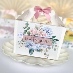 Peut inclure: Boîte à dragées blanche avec un ruban rose et un motif floral aquarelle. La boîte a une poignée et indique "The Wedding of Lewis & Nicole 01.07.2024".