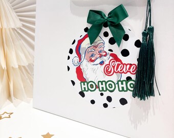 May include: White gift bag with a Santa Claus graphic on a black polka dot background. The graphic includes the name "Steve" and the phrase "HO HO HO". A green bow and tassel add a festive touch to the bag.