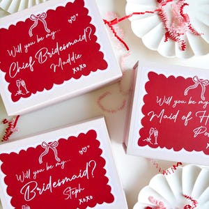 May include: Three pink gift boxes with red and white labels. The labels have a white scalloped border and say "Will you be my... Chief Bridesmaid? Maddie xoxo", "Will you be my... Bridesmaid? Steph xoxo", and "Will you be my Maid of Honor?"